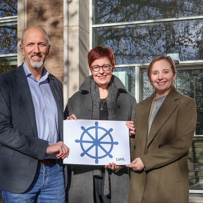 Drei Menschen halten ein Bild mit einem Steuerrad hoch (vergrößerte Bildansicht wird geöffnet)
